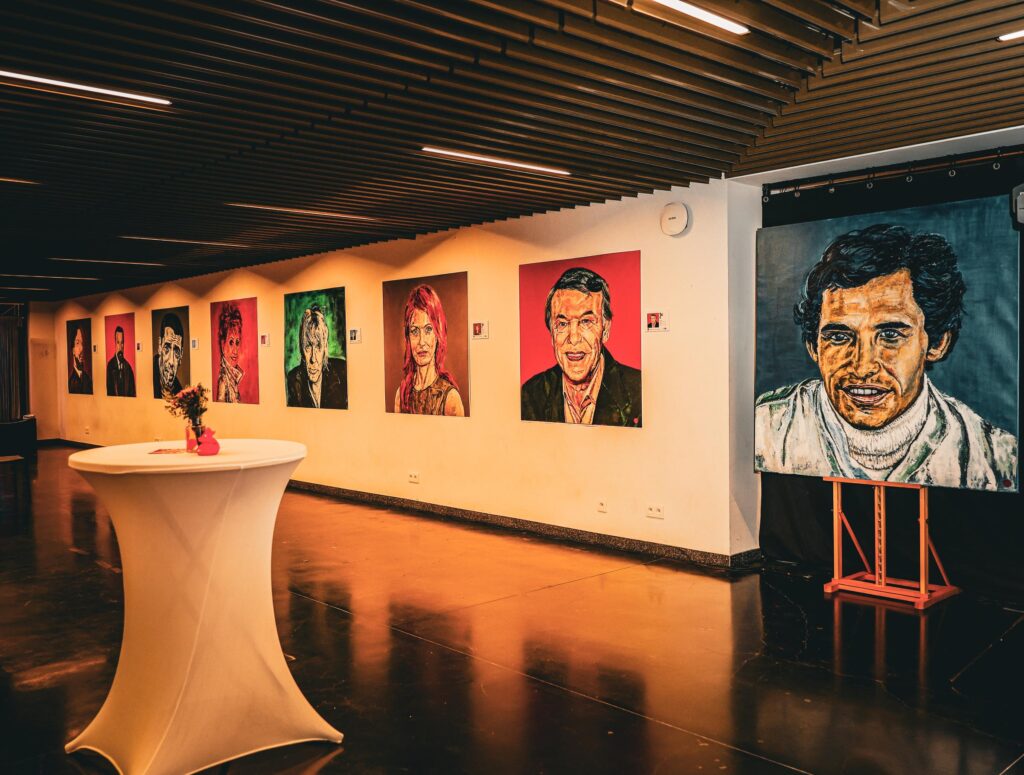 Exhibition venue in Brussels with large painted portraits on the walls, a cocktail table and a portrait on an easel