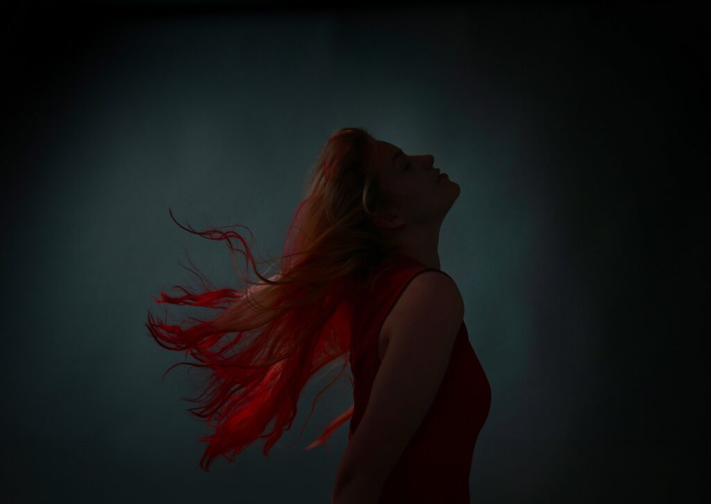 Fine art silhouette portrait of a woman in profile, head tilted back, red-lit hair streaming against a dark teal backdrop