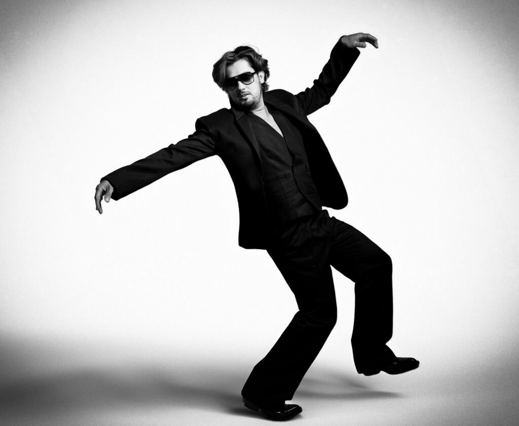 Black and white fine art portrait of a man in a dark suit and sunglasses striking a playful dance pose, white studio