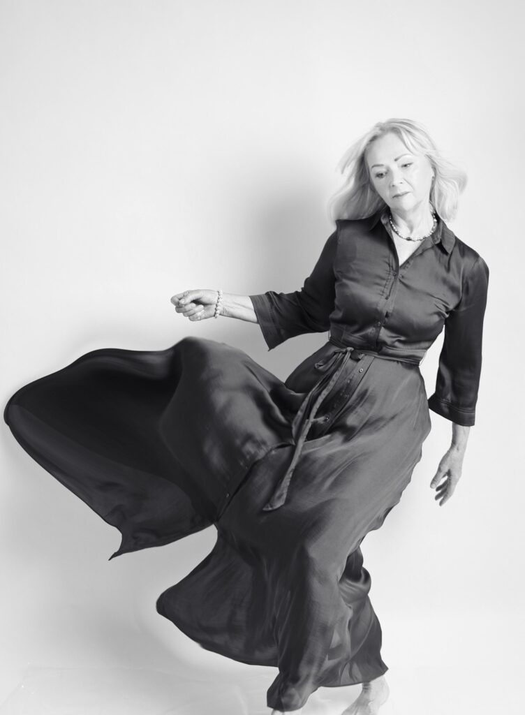 Black and white fine art portrait of a mature woman twirling in a flowing satin shirt dress, pearls, high-key studio