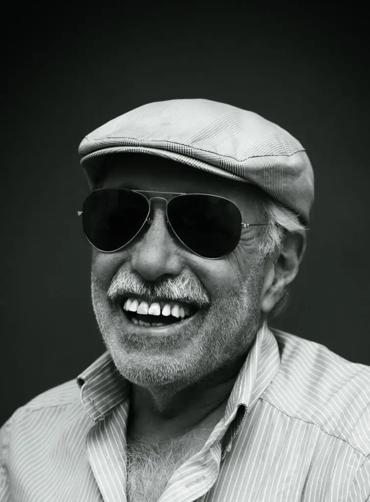 Black and white studio portrait of a laughing man in a flat cap and aviator sunglasses, Brussels