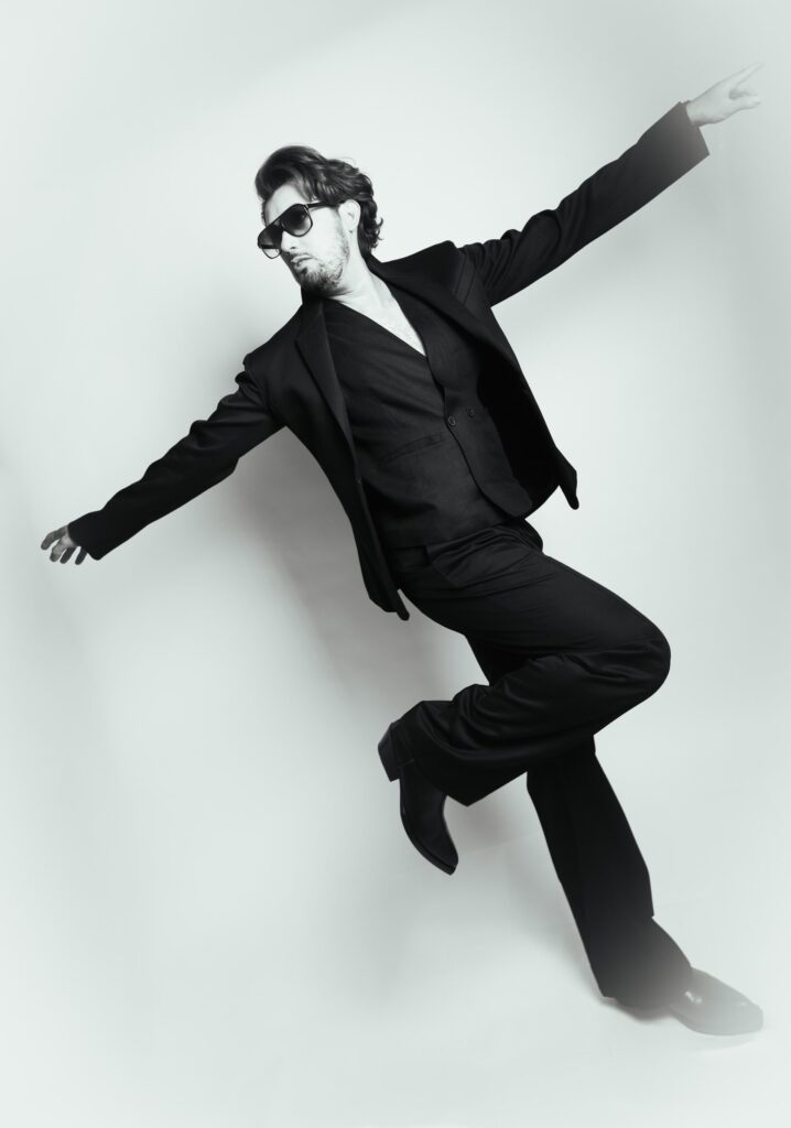 Black and white fine art portrait of a man in a black suit leaping mid-dance, arms outstretched, light studio