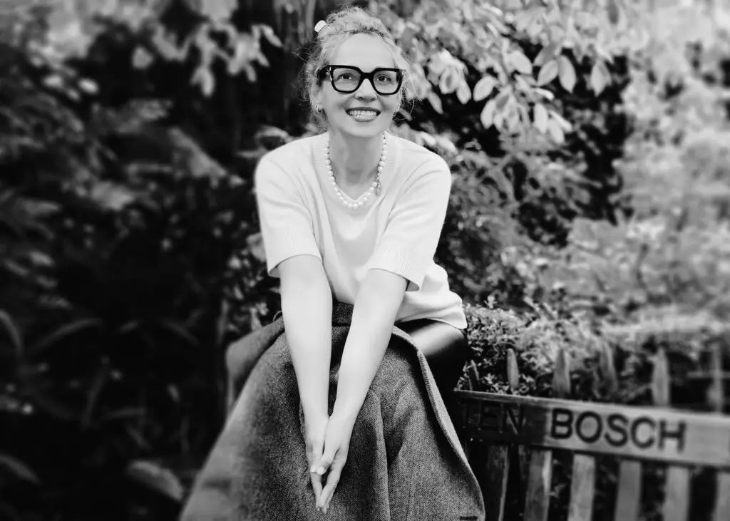 Black and white outdoor portrait of a smiling woman in bold glasses and pearls on a bench in Tenbosch Park, Brussels
