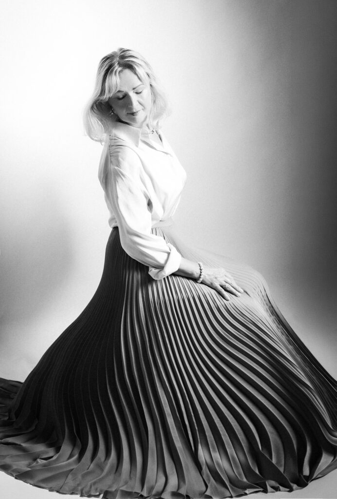 Black and white fine art portrait of a woman seated in a flowing pleated skirt and white blouse, eyes closed, soft light