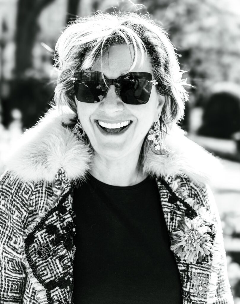 Black and white outdoor portrait of a laughing woman in oversized sunglasses and a fur-collared tweed jacket, Brussels