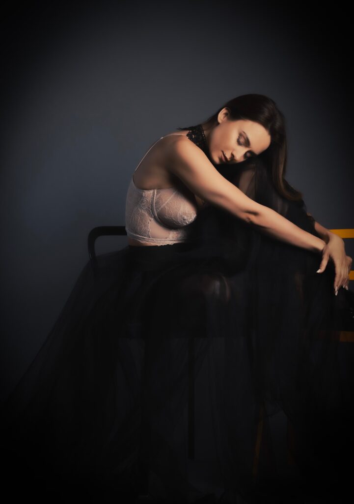 Boudoir portrait of a woman in a lace top and black tulle skirt, seated with eyes closed against a dark studio backdrop in Brussels