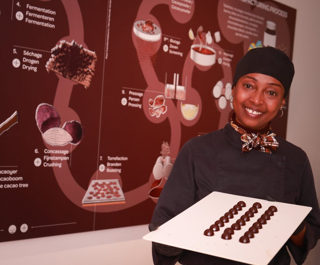 Smiling chocolatier presenting a tray of handmade pralines in front of a cocoa-process wall, Brussels