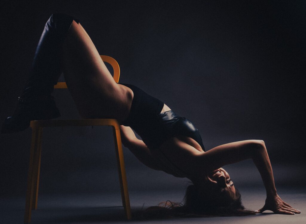Creative boudoir photo of a laughing woman in a black bodysuit and boots, arching backwards over a yellow chair