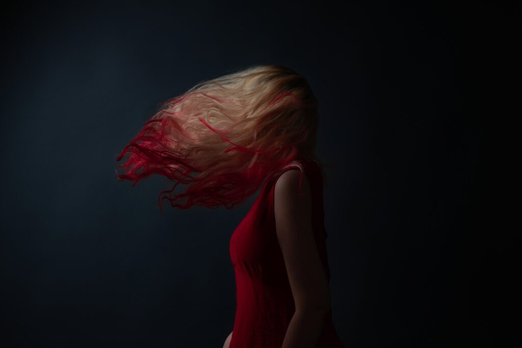 Fine art portrait of a woman in a red dress tossing her red-tipped blonde hair across her face, dark studio background