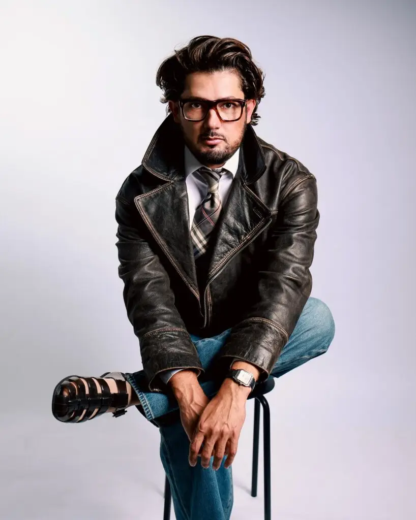Editorial portrait of a man in a leather jacket, tie and jeans seated on a stool, direct gaze, light backdrop
