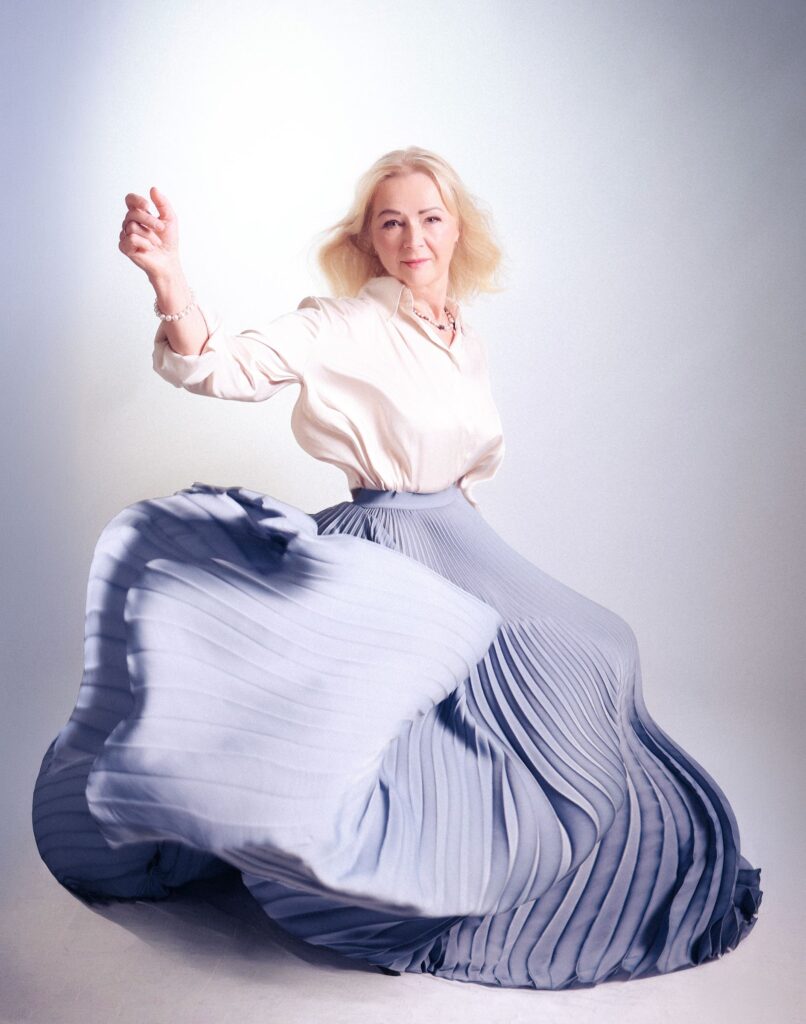 Fine art portrait of a mature woman in a white satin blouse twirling a pale blue pleated skirt, pearls, airy light