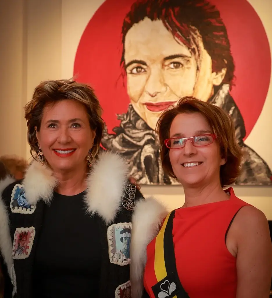 Two smiling guests posing in front of a large painted portrait on a red background at an exhibition opening in Brussels