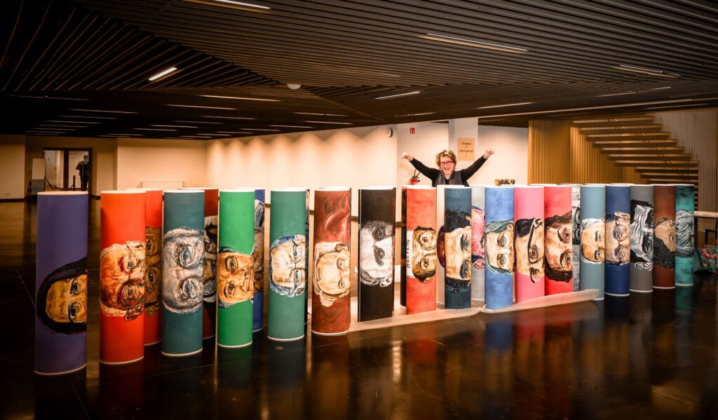 Artist celebrating with raised arms behind a row of rolled painted portraits while setting up an exhibition in Brussels