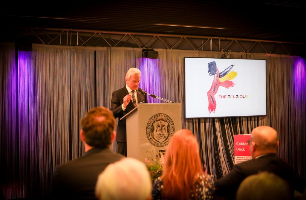 Speaker at a City of Brussels podium during The Belgious exhibition opening, guests listening in the foreground
