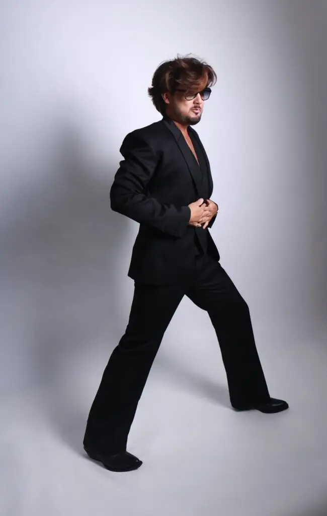 Fashion portrait of a man in a black suit and sunglasses striding across a light studio backdrop