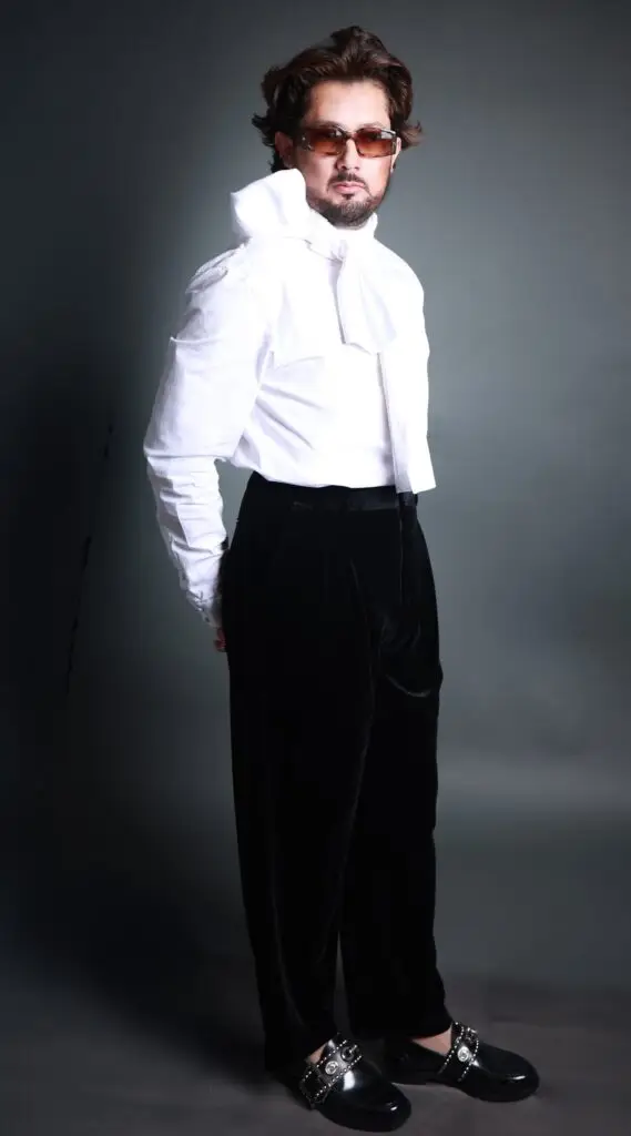 Full-length fashion portrait of a man in a white tie-neck shirt, black velvet trousers and tinted sunglasses