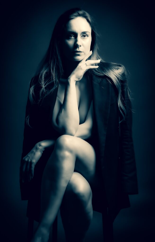 Fine art boudoir portrait of a confident woman seated with crossed legs, a dark blazer over her shoulders, cool-toned studio light