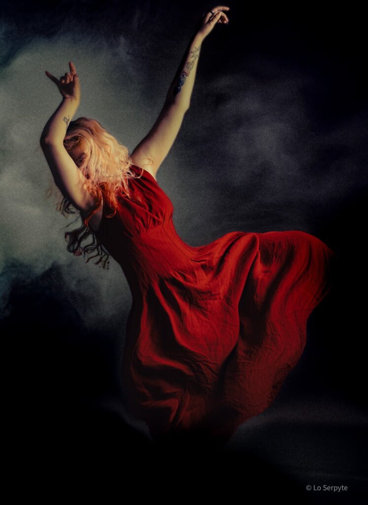 Fine art portrait of a woman dancing in a flowing red dress, arms raised against a dark smoky sky, painterly style