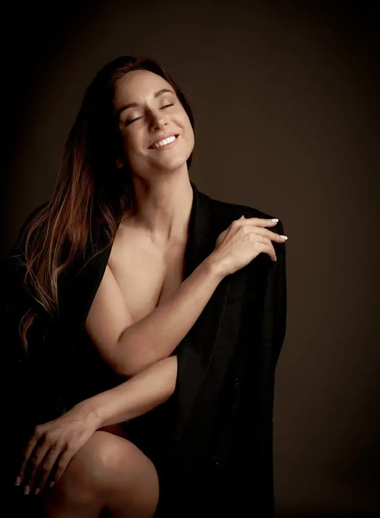 Joyful boudoir portrait of a smiling woman with eyes closed, wrapped in a black blazer, warm brown backdrop