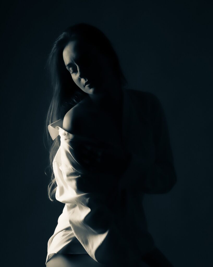 ow-key boudoir portrait of a woman in a white shirt emerging from the shadows, looking down, Lo Portraits Brussels