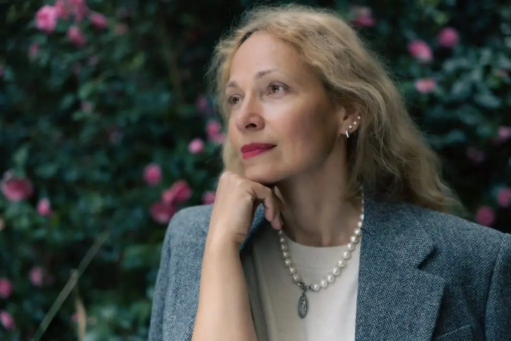 Thoughtful outdoor portrait of a woman in pearls and a grey blazer, chin resting on her hand, in front of pink roses
