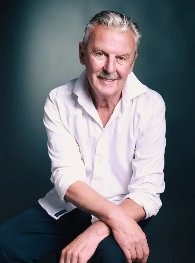 Warm studio portrait of a smiling mature man in a white shirt, arms resting on his knee, teal backdrop