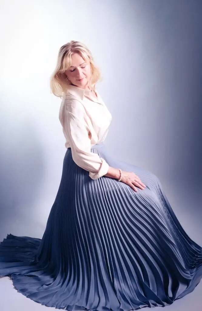 Soft studio portrait of a woman seated in a sweeping blue pleated skirt, eyes closed, gentle smile