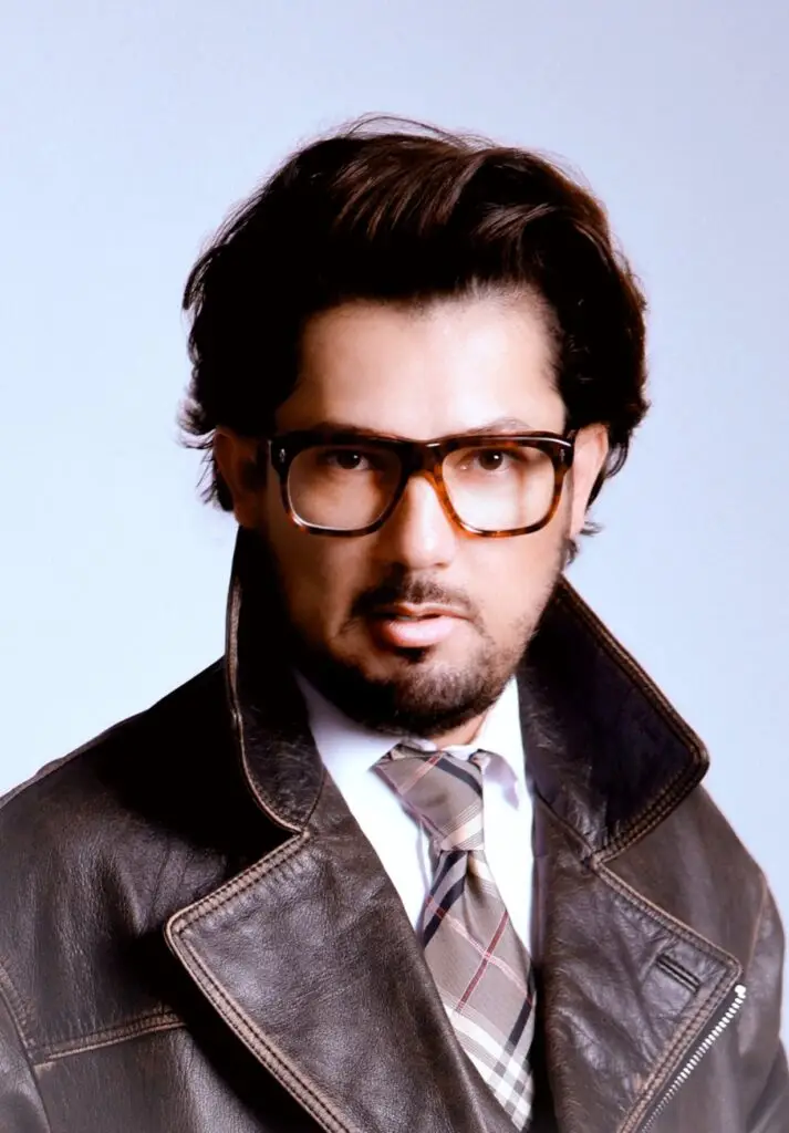 Studio portrait of a bearded man in tortoiseshell glasses, leather jacket and plaid tie
