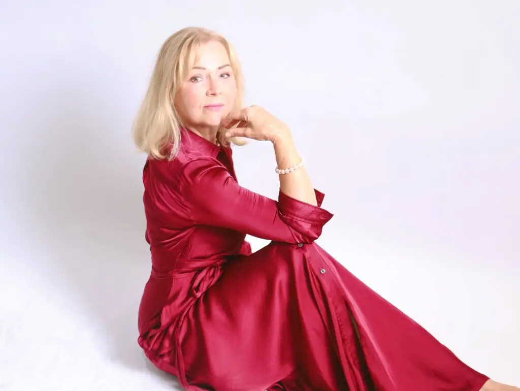 Studio portrait of a woman in a burgundy satin dress seated on the floor, chin resting on her hand, pearl bracelet