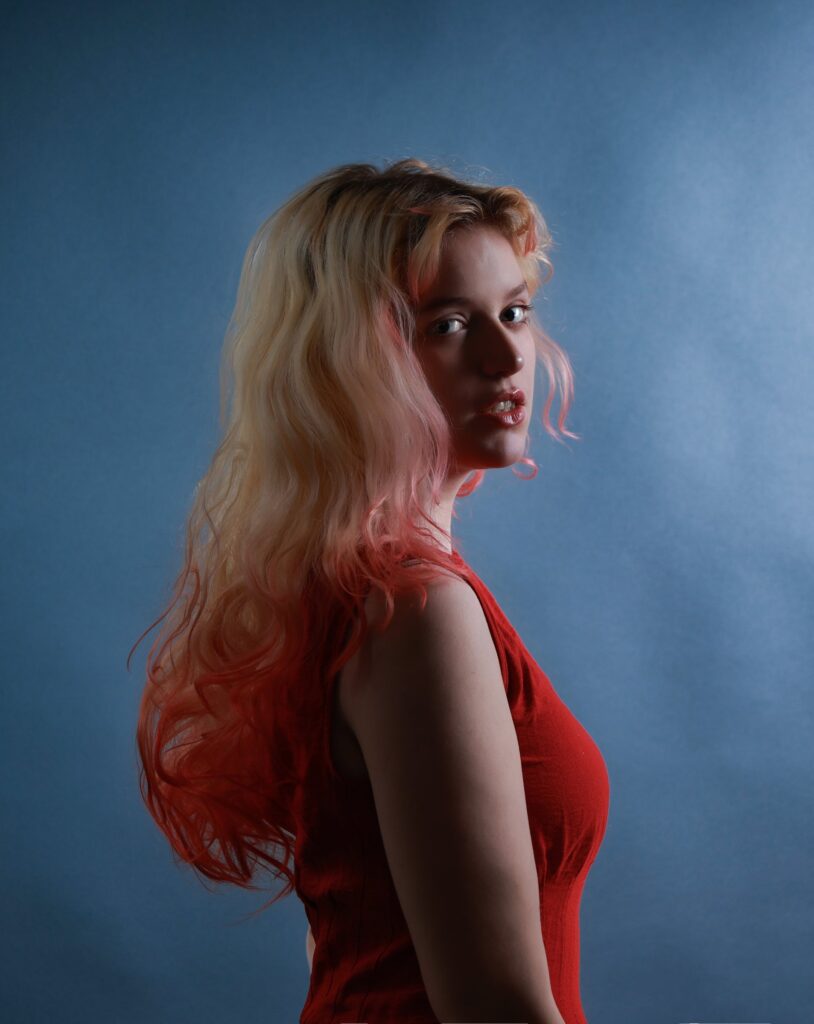 Portrait of a young woman in a red top with pink-tipped blonde hair looking over her shoulder, blue studio backdrop